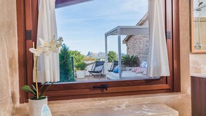 Ein Badezimmer mit Badewanne, einem Fenster mit Vorhängen und Blick auf Sitzgelegenheiten und Pflanzen im Freien.
