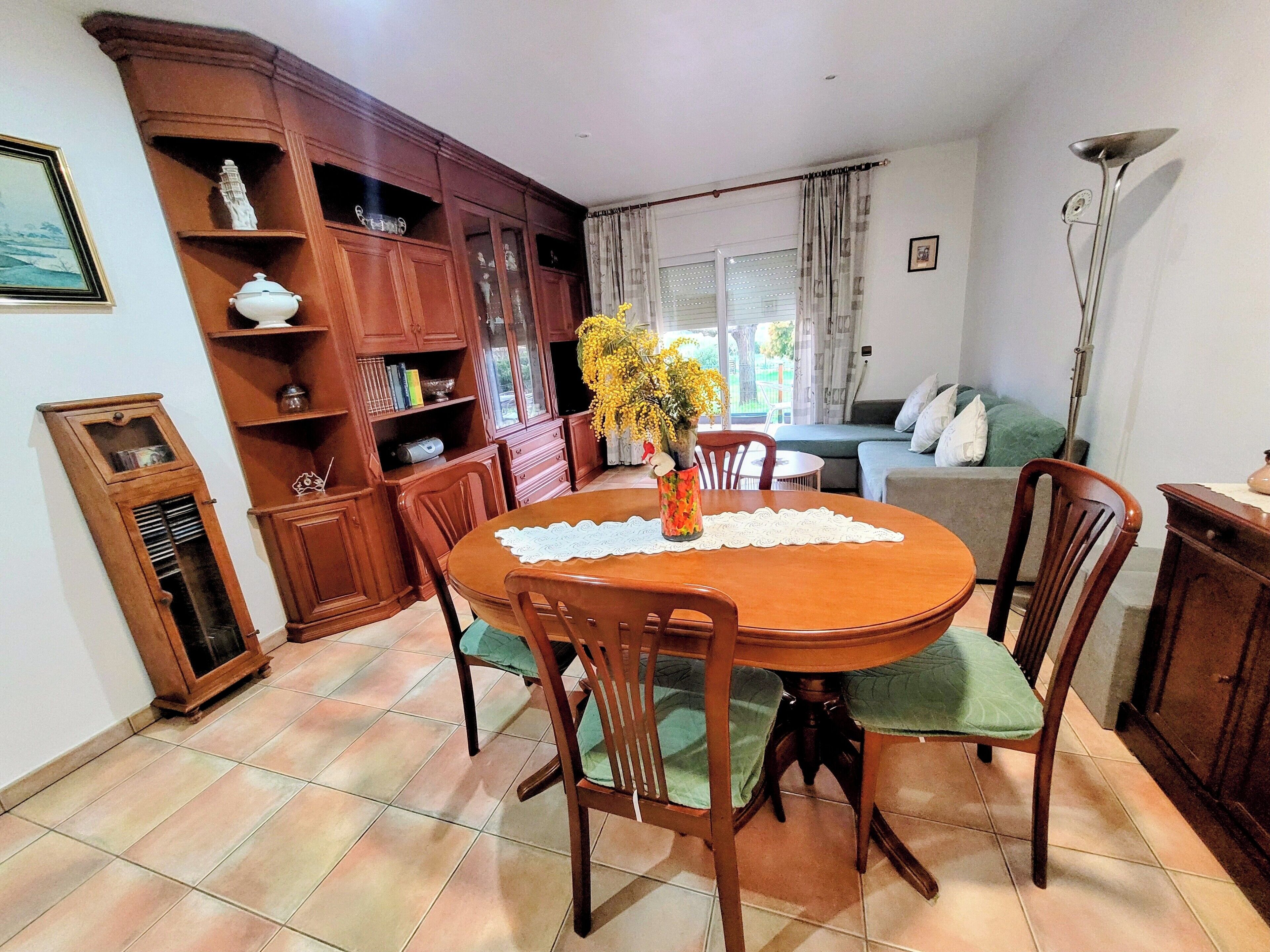 A dining area with a wooden table, chairs, and a vase of flowers. There is a cabinet with shelves and a television in the background.