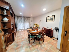 A dining area with a wooden table, chairs, and a vase of flowers. There is a bookshelf with decorative items and a picture on the wall.