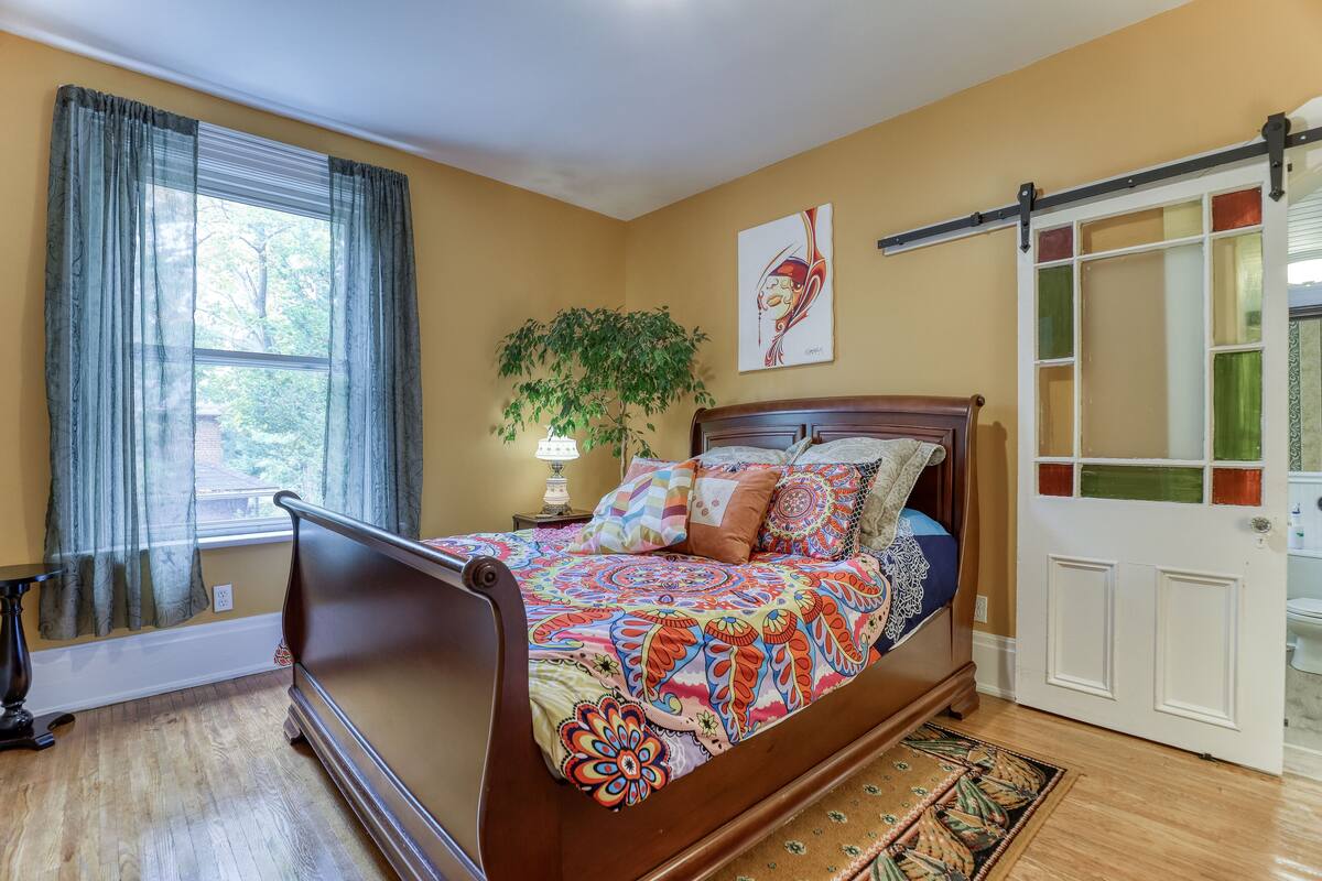 A bedroom with a wooden bed, a large window with curtains, a potted plant, and a door with a glass panel.