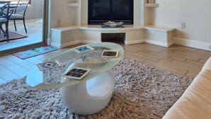 Een woonkamer met een open haard, een flatscreen-tv erboven en een glazen salontafel met decoratieve objecten.