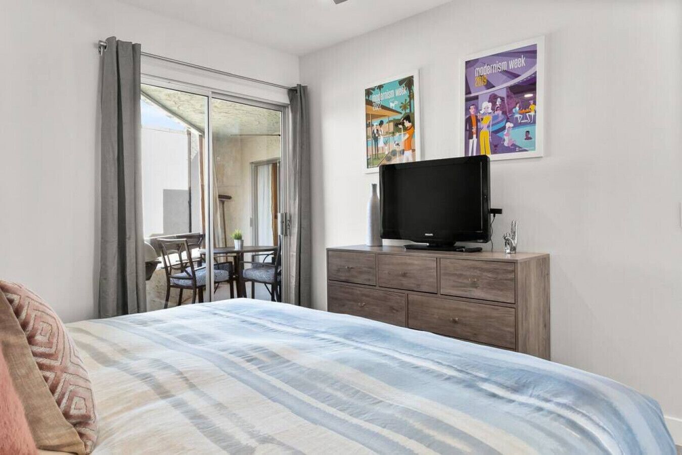 A bedroom with a bed, a TV on a wooden dresser, a dining area with chairs and a table, and two posters on the wall.