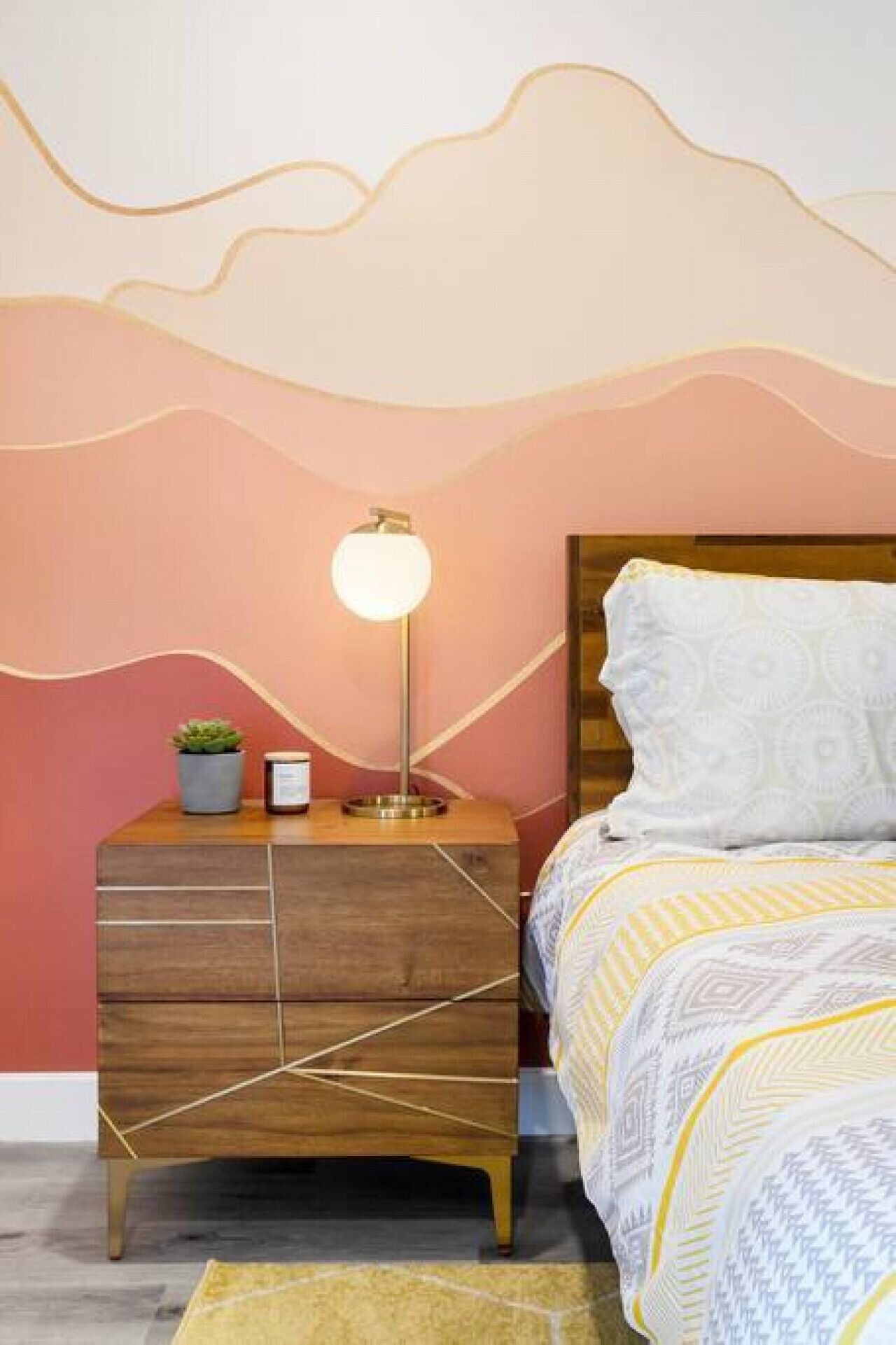 Quarto com mesa de cabeceira de madeira, luminária, planta pequena e colcha estampada.