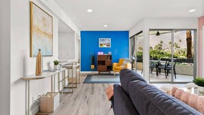 Ein modernes Wohnzimmer mit blauer Akzentwand, einer Couch, einem Essbereich mit Tisch und Stühlen sowie einem Gartenblick durch große Fenster.