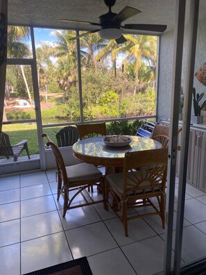 Eine überdachte Terrasse mit Tisch und Stühlen, große Fenster mit Blick auf Palmen und ein Deckenventilator.