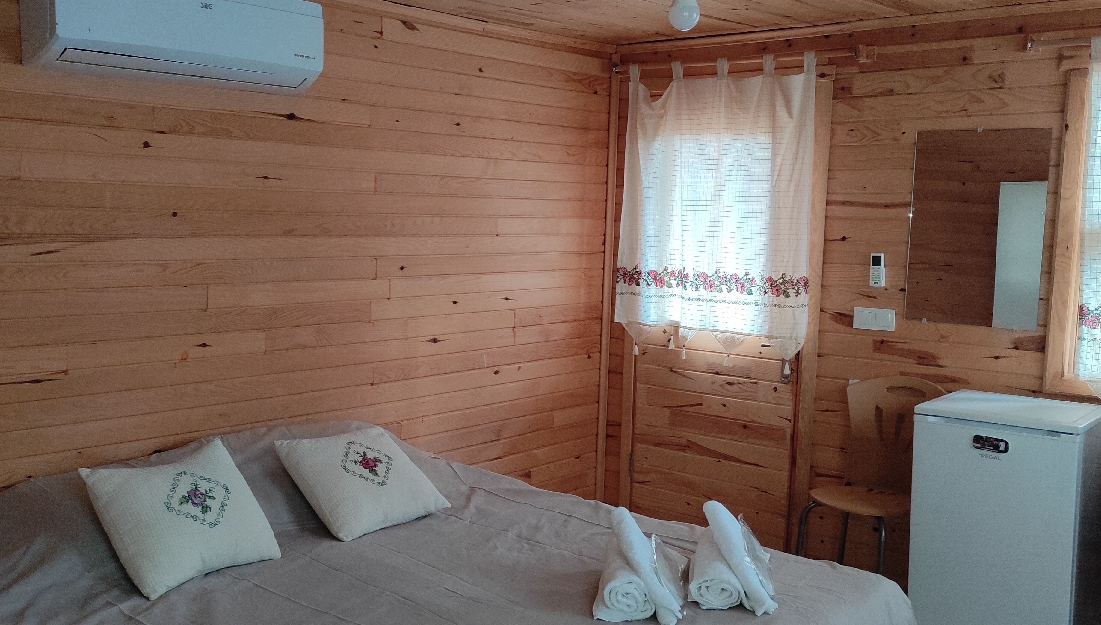 A wooden cabin room with a bed, two pillows, a small table with towels, a mirror, and a white refrigerator.