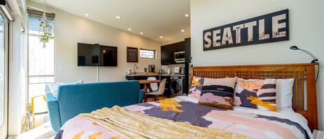 A modern hotel room with a bed, a sofa, a TV, a kitchen area, and a large 'SEATTLE' sign on the wall.