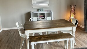 Un comedor con mesa de madera y bancos, una silla blanca, un estante pequeño con libros y una lámpara decorativa.