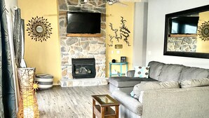 Una sala de estar con chimenea de piedra, un sofá gris, una mesa de centro de madera y un televisor montado en la pared.