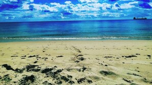 En strand med fodspor, der fører hen til vandet, et skib i det fjerne og en klar himmel.