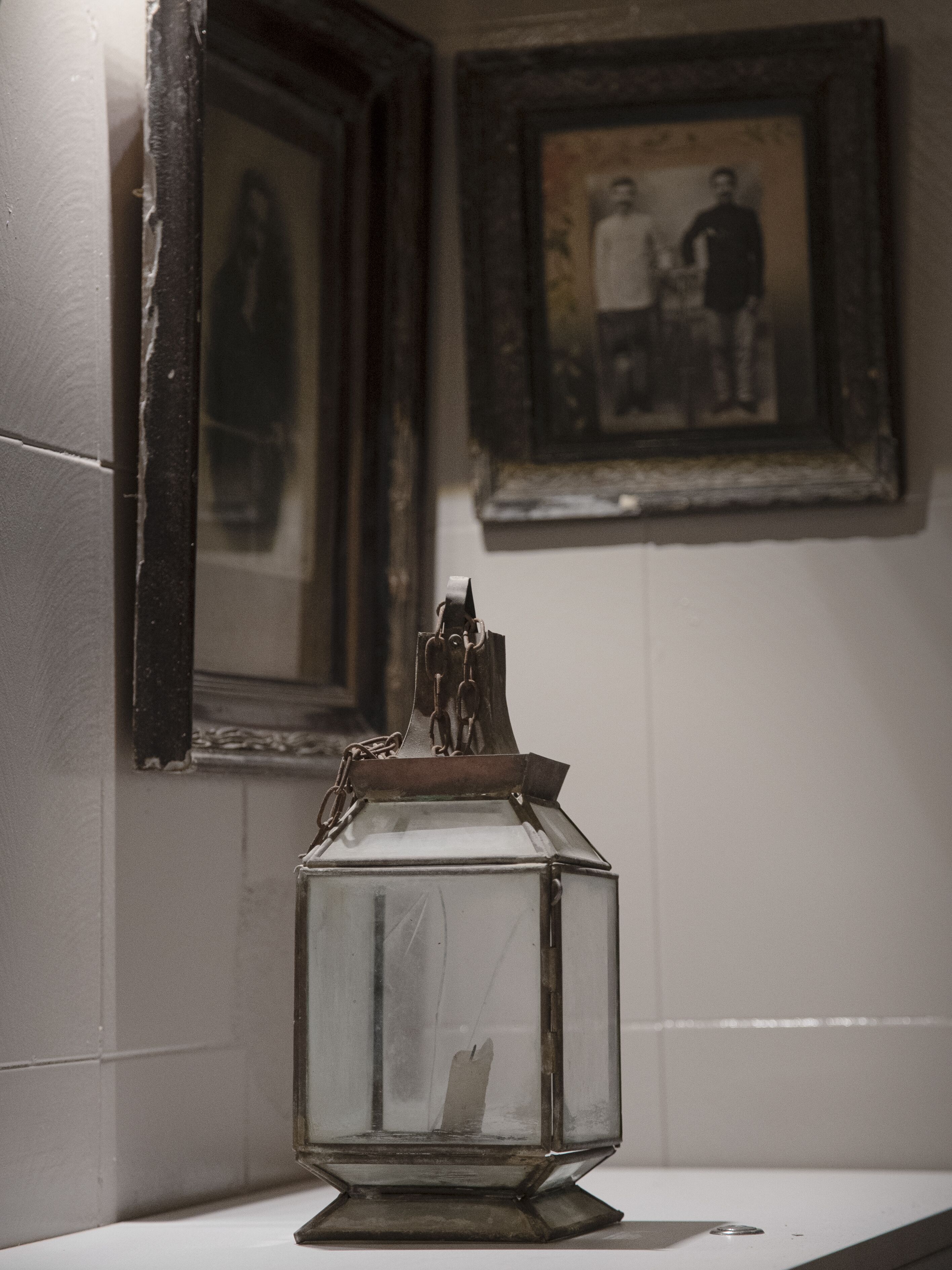 A glass lantern on a white surface with two framed pictures on the wall behind it.