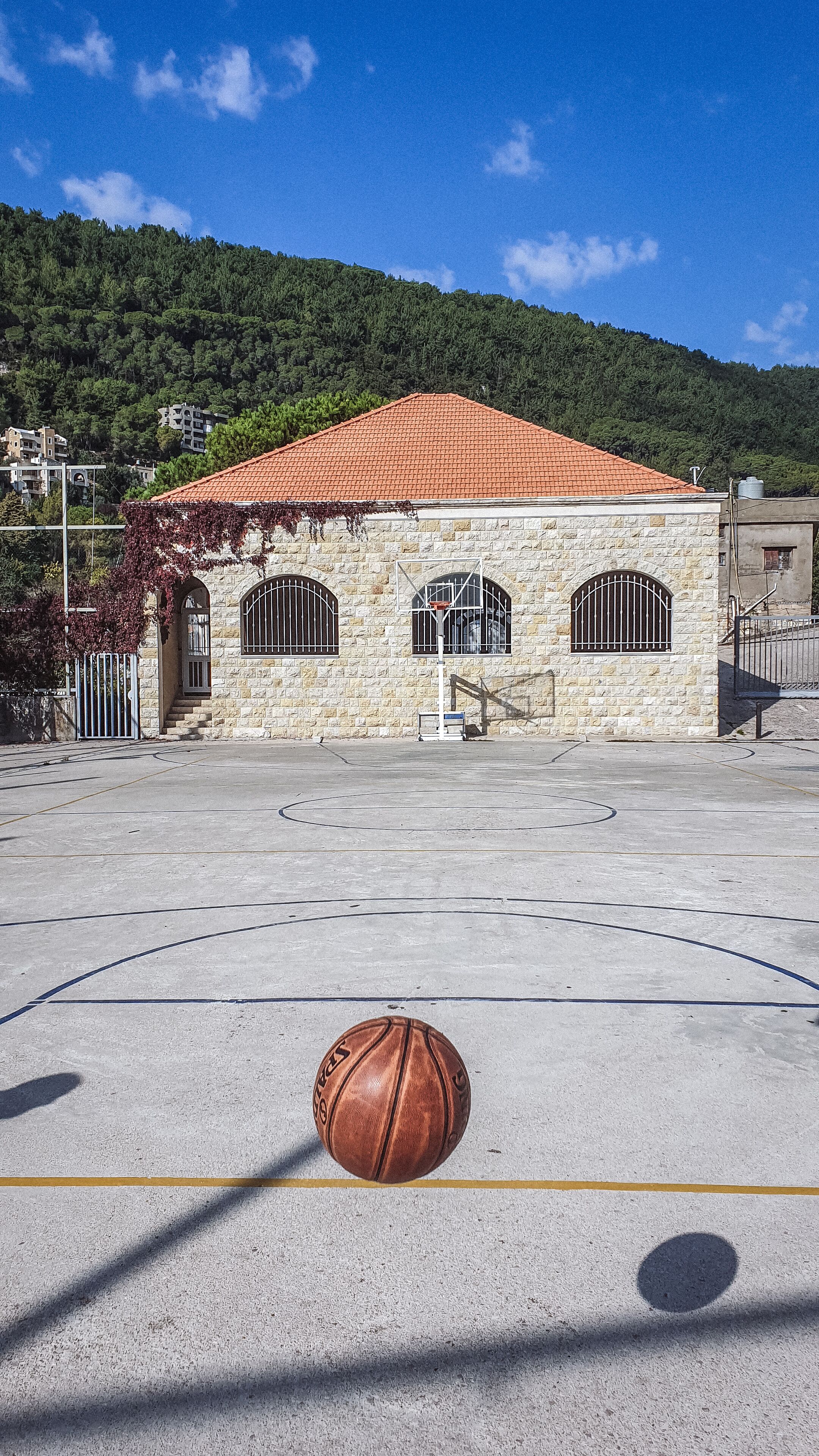 A basketball court with a ball in the foreground, a building with a red-tiled roof, and a forested hillside in the background.