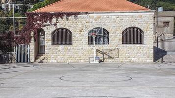 A basketball court with a ball in the foreground, a building with a red-tiled roof, and a forested hillside in the background.
