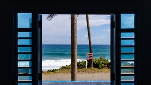 A view through a window with blinds, featuring a beach with palm trees and a flag. - breathtaking sea view Beach FRONT very PRIVATE and quiet SOLAR POWERED (Arecibo)
