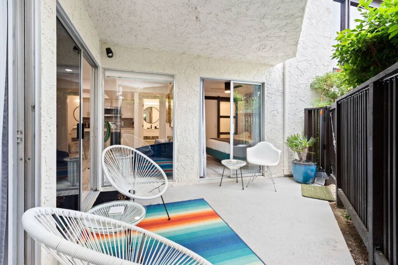 Eine moderne Terrasse mit weißen Stühlen, einem bunten Teppich und einer Glastür zu einem Innenraum.