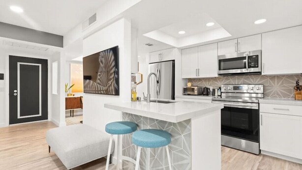 A modern kitchen with white cabinetry, a central island with bar stools, stainless steel appliances, and a geometric backsplash. - Newly renovated, vibrant and chic Biltmore condo (Palm Springs)
