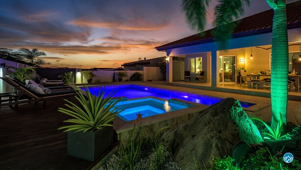 Una piscina con vistas al atardecer, un patio con sillones de descanso y una casa bien iluminada con comedor.