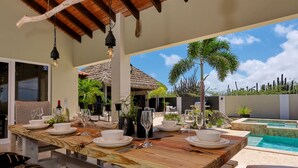 Un área de comedor al aire libre con una mesa de madera preparada para una comida, rodeada de plantas en macetas y con vistas a una piscina.