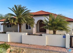 Une maison moderne avec un toit recouvert de tuiles rouges, une grande porte d’entrée en arc de cercle et un jardin bien entretenu, agrémenté de palmiers et d’arbustes.