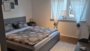 Un dormitorio moderno con una cama grande, una mesita de noche, un escritorio y una ventana con cortinas traslúcidas.