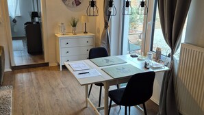 Una habitación con suelo de madera, un escritorio blanco con papeles, una silla negra, un reloj y una ventana con cortinas.