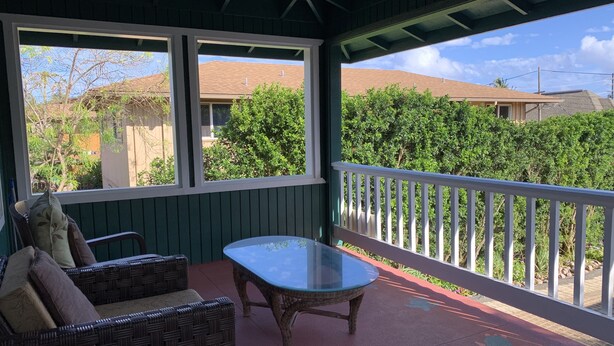 Eine überdachte Veranda mit Korbmöbeln, einem Tisch mit Glasplatte und Blick auf Nachbarhäuser und Grünflächen.