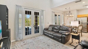 A spacious living room with a leather sofa, a dining table with chairs, a chandelier, and a refrigerator. - The Secret Serenity Villa – 3 Miles from Disney, Endless Comfort & Fun (Kissimmee)