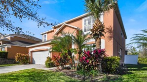 A two-story house with a garage, surrounded by well-maintained landscaping including palm trees and colorful plants. - The Secret Serenity Villa – 3 Miles from Disney, Endless Comfort & Fun (Kissimmee)