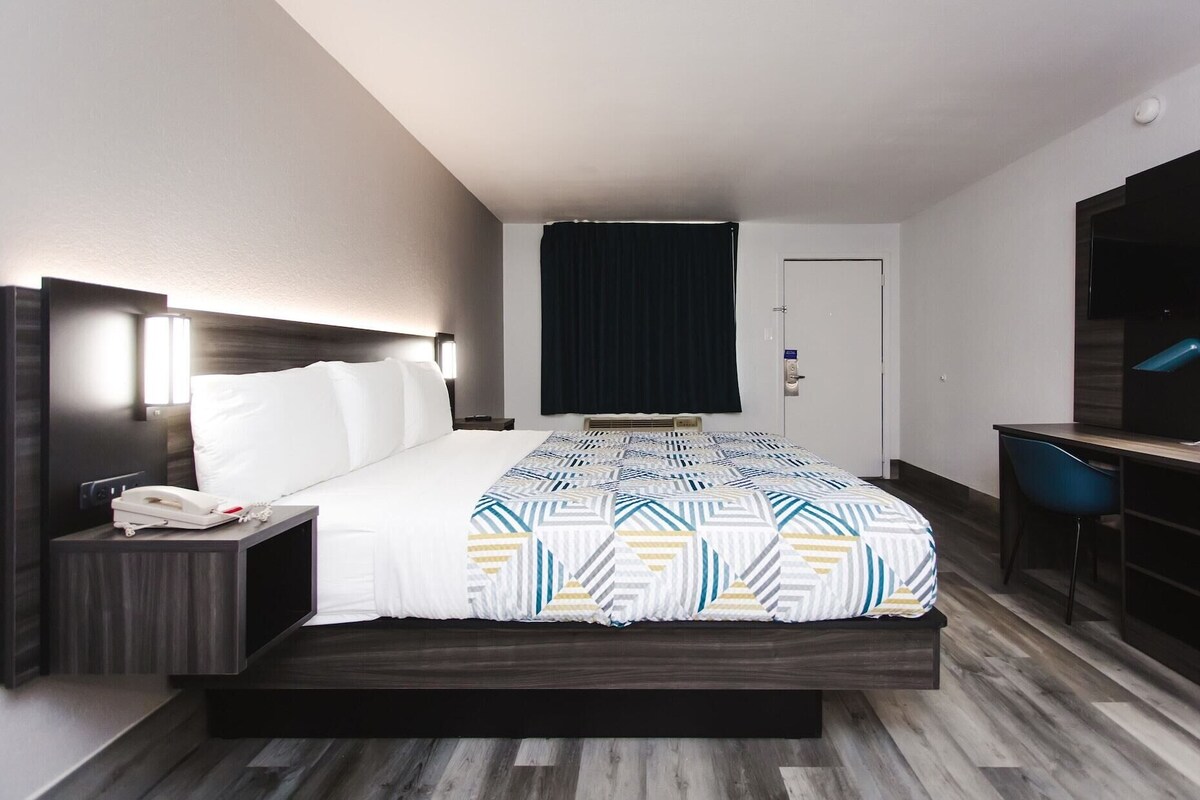 Hotellihuone, jossa on sänky, yöpöytä puhelimella, työpöytä tuolilla ja tumma verho.