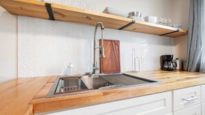 Una cucina con un piano di lavoro in legno, un lavello in acciaio inossidabile e un forno integrato. Sopra il lavello, mensole in legno con stoviglie e bicchieri.