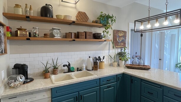 Een keuken met blauwgroene kastjes, een wit aanrecht en houten vloeren. Er hangen lampen, er staat een koffiezetapparaat en er staan diverse keukenbenodigdheden op de planken.