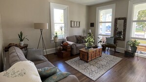 Een ruime woonkamer met een grijze bank, een salontafel en een fauteuil. Er staan potplanten en er is een vloerlamp.