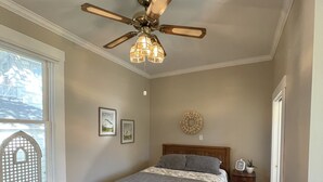 Een slaapkamer met een houten vloer, een bed met een grijze dekbedovertrek, een plafondventilator en een raam met een wit traliewerk.