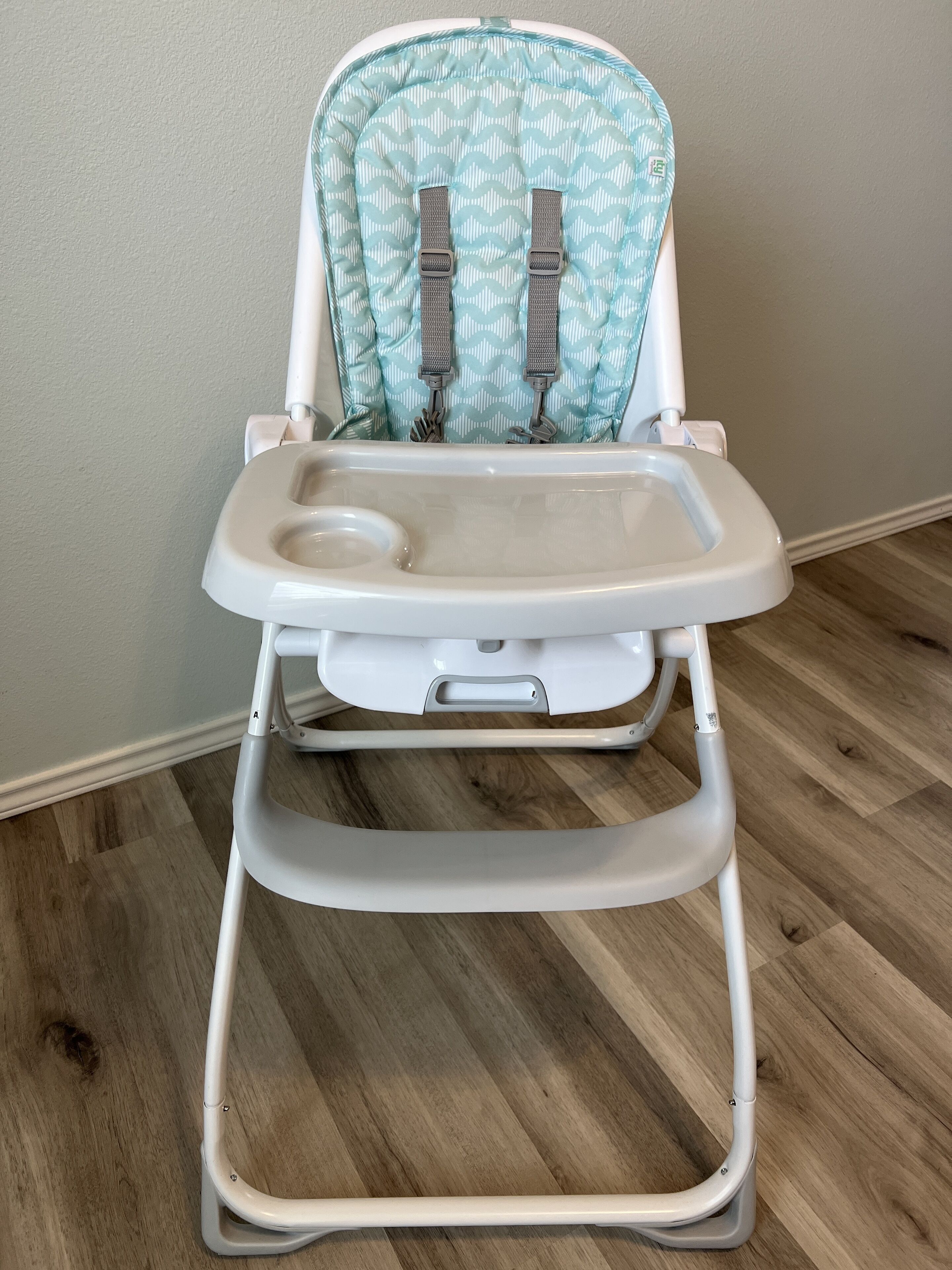 A white high chair with a patterned seat and a tray, placed on a wooden floor against a plain wall.