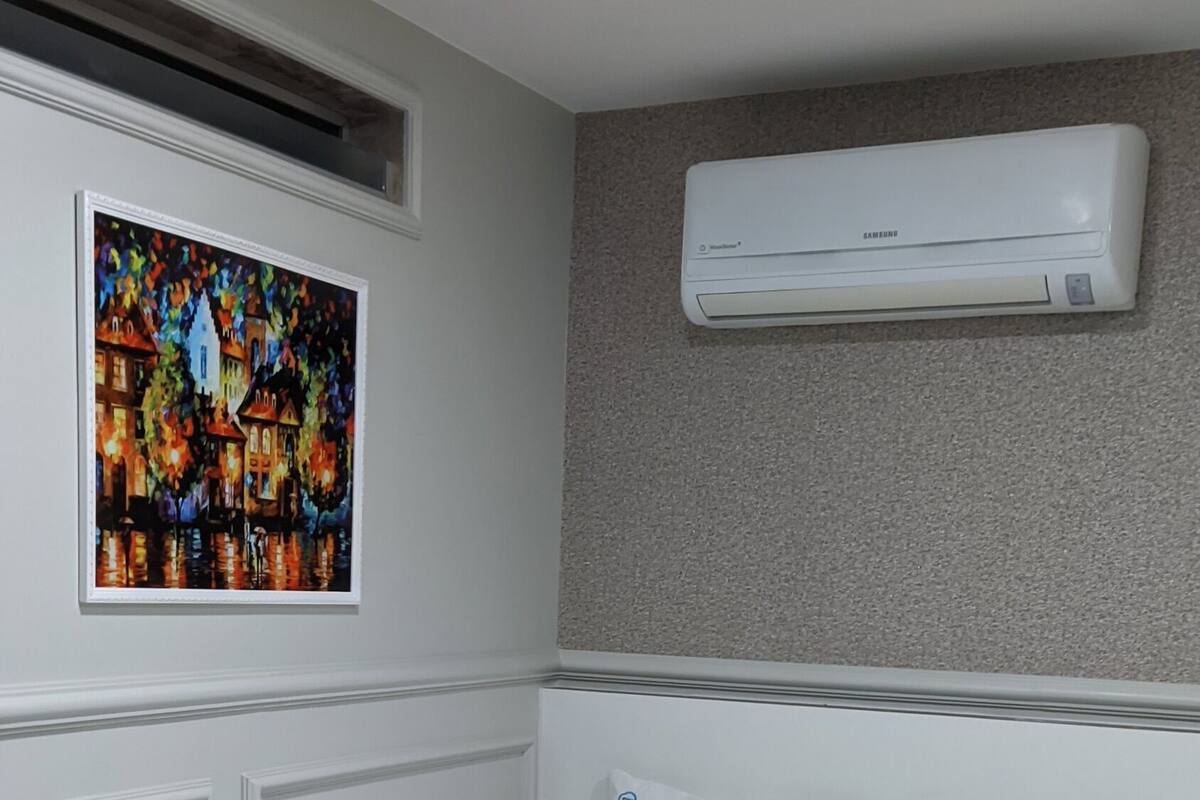 Habitación de hotel con cama, ropa de cama blanca, una manta color turquesa, unidad de aire acondicionado y un cuadro en la pared.