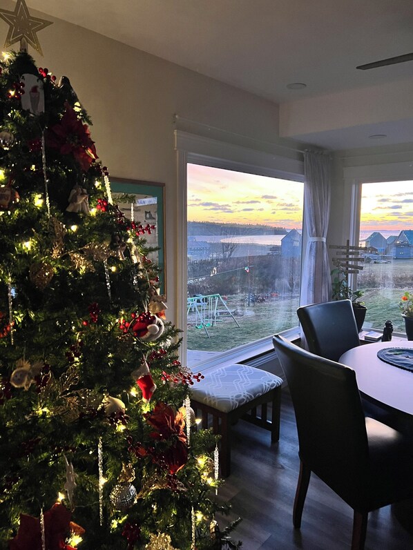 A room with a Christmas tree, a dining table with chairs, and a view of a snowy landscape through large windows.