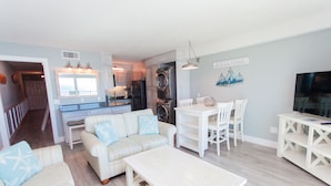 A modern living room with a sofa, coffee table, dining table, and chairs. There is a kitchen area with appliances and a dining area with a TV. - Beautiful Gulf Front Condo!  Summit 627 (Panama City)