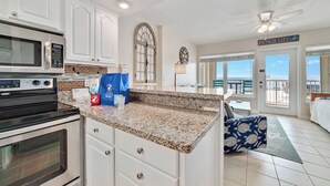 Eine Küche mit weißen Schränken, einer Granit Arbeitsplatte, einem Edelstahlbackofen und einer Mikrowelle. Ein Fenster bietet Blick auf den Strand.