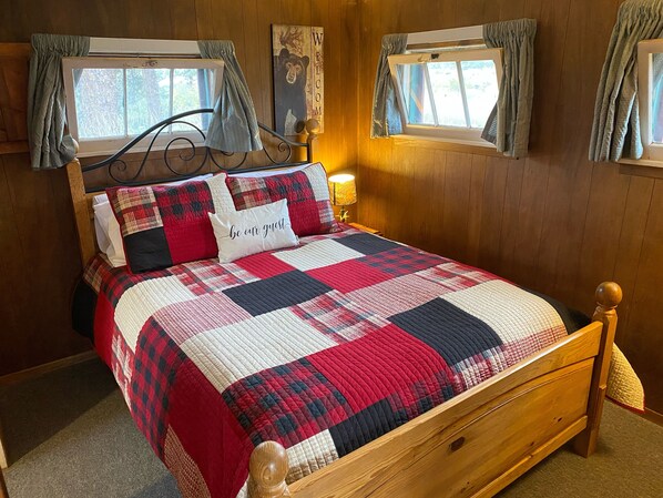 A wooden bed with a red and black quilt, a pillow with "be our guest," and a "welcome" sign above the window. - Marr's Mountain Cabins (Red Feather Lakes)