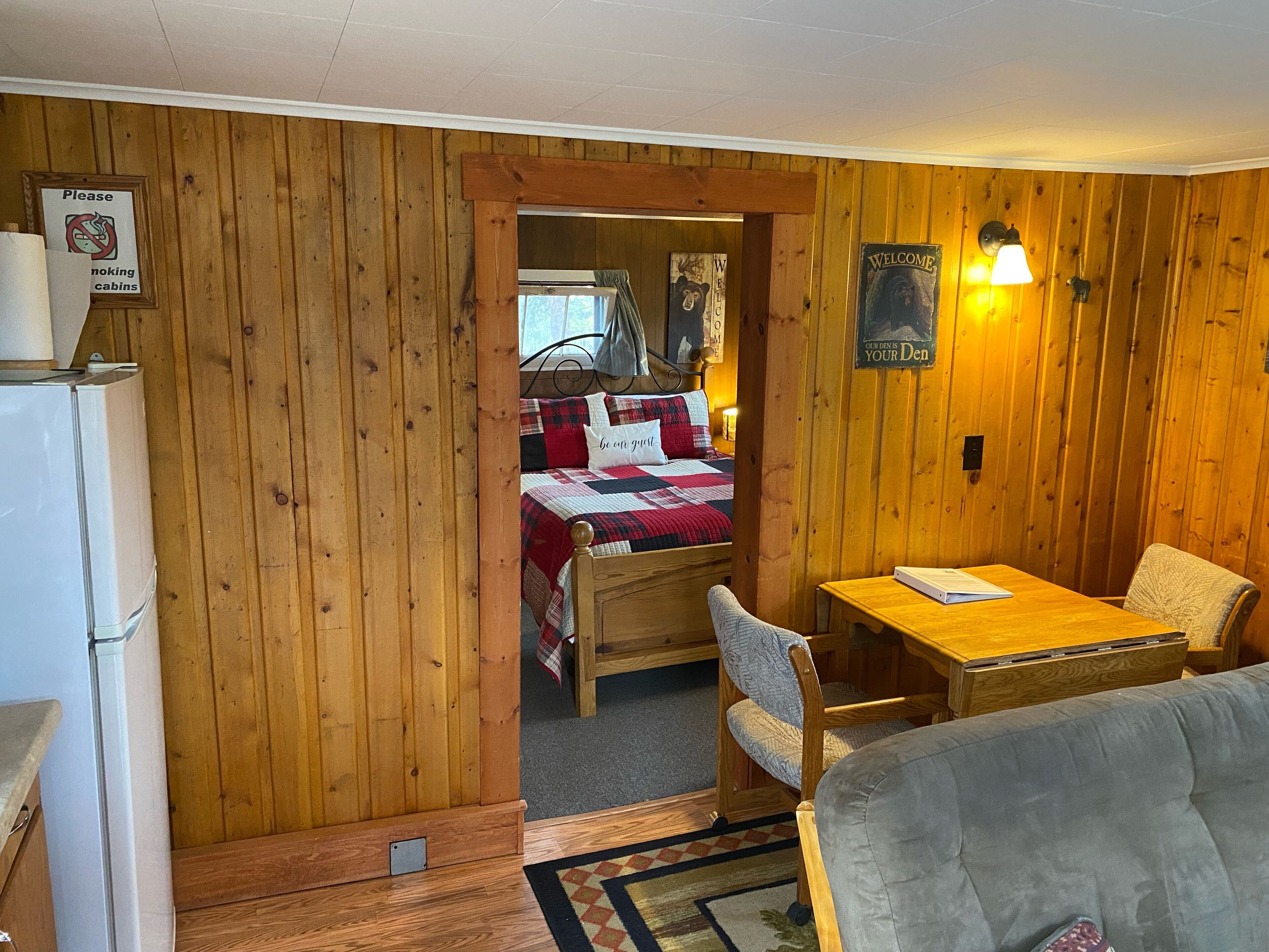 A cozy cabin-style room with wooden walls, a bed with a red and white quilt, a small table with a chair, and a refrigerator.