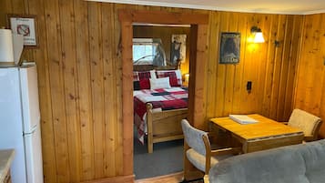 A cozy cabin-style room with wooden walls, a bed with a red and white quilt, a small table with a chair, and a refrigerator.