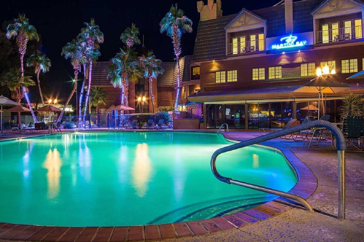 A hotel with a swimming pool, palm trees, and a well-lit exterior.