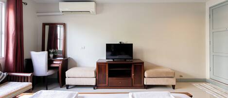 A hotel room with a bed, two armchairs, a desk with a television, a mirror, and an air conditioning unit.