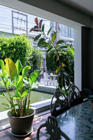 Une vue depuis une fenêtre, avec une plante en pot au premier plan, une petite table avec des chaises, et un bâtiment avec des balcons en arrière-plan.
