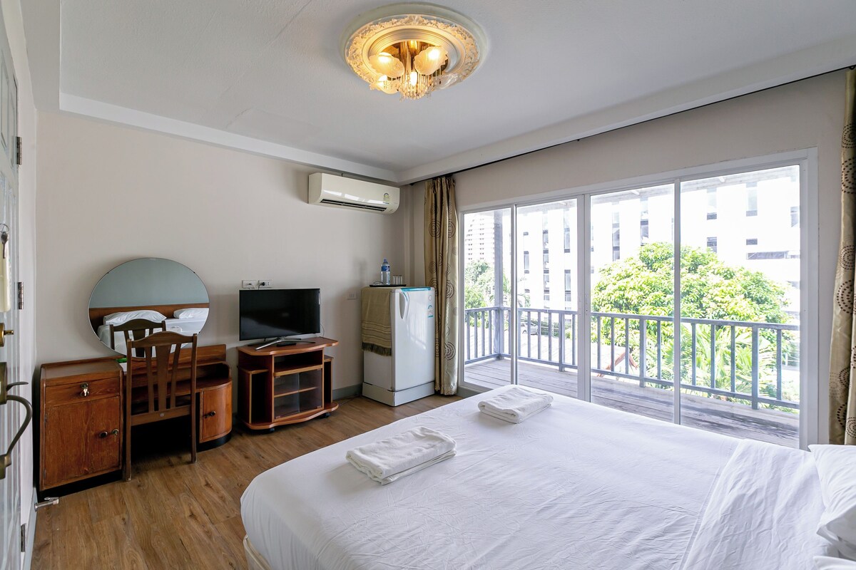 Habitación de hotel con cama, escritorio con televisor, armario, equipo de aire acondicionado y balcón con vista a edificios y follaje.