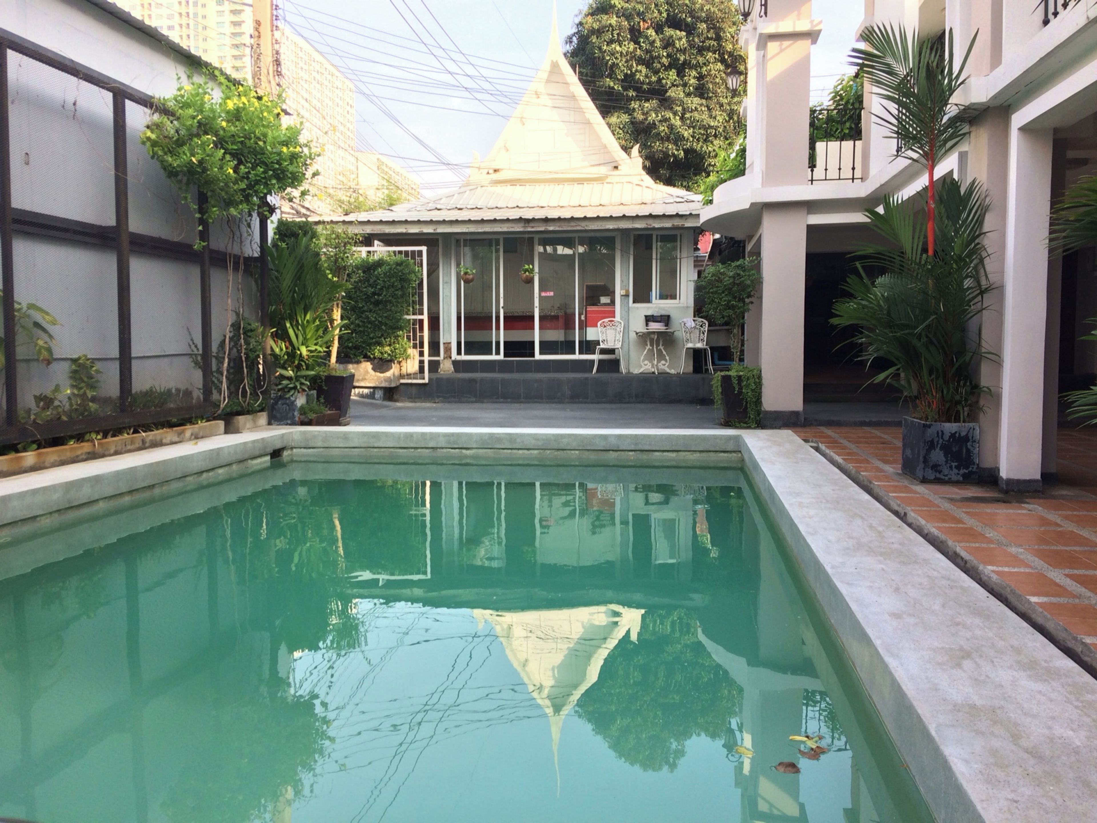 A swimming pool with a tiled deck, surrounded by potted plants and a building with a traditional roof.