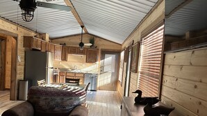A spacious cabin interior with a wooden floor, a dining area, a kitchen, and a large window allowing natural light.