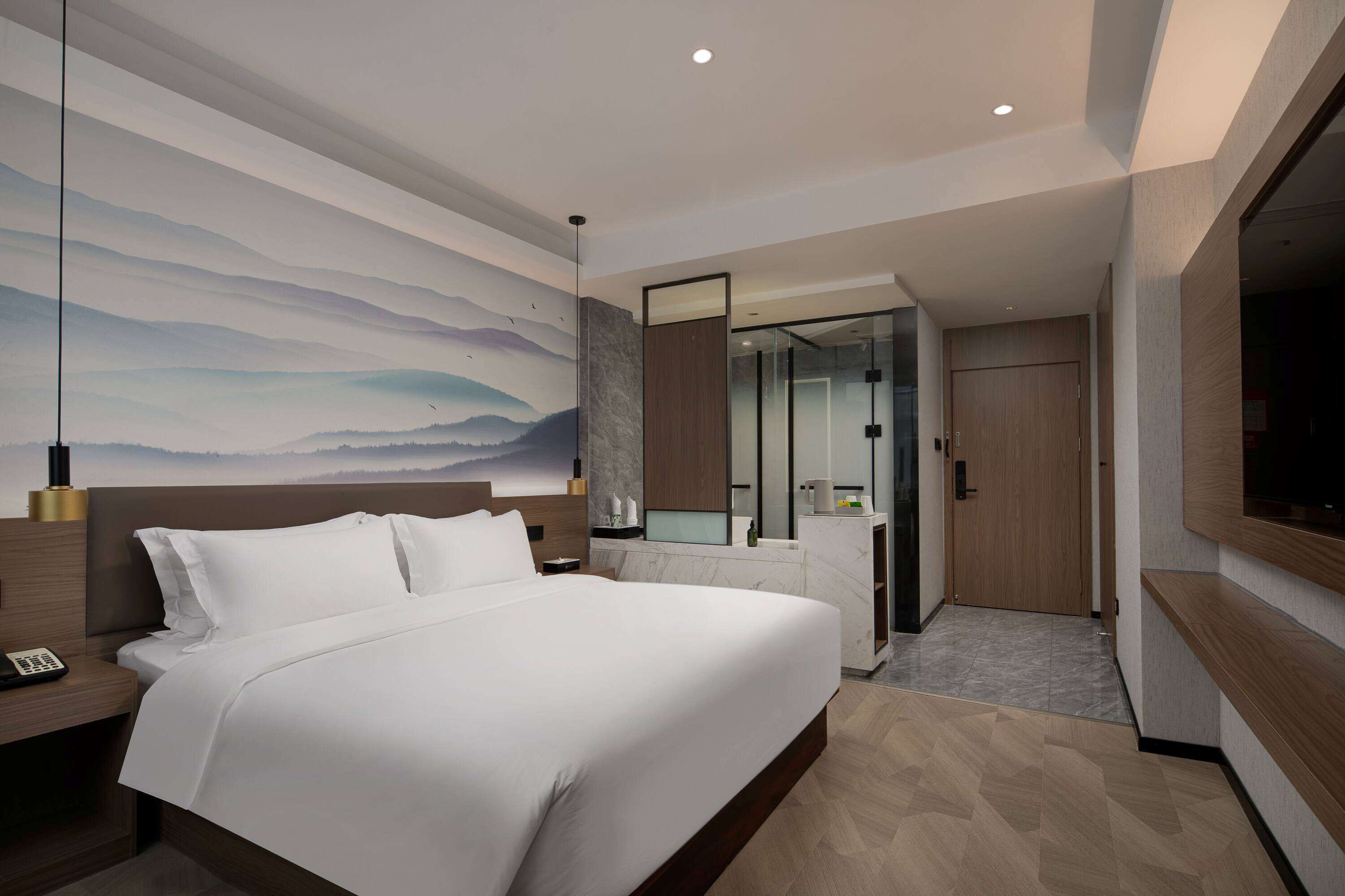 A modern hotel room with a large bed, a nightstand, a wall-mounted TV, and a bathroom with a glass partition.