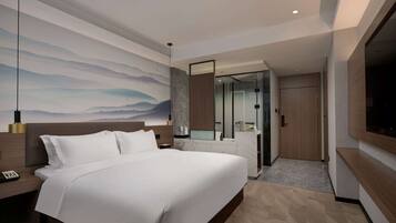 A modern hotel room with a large bed, a nightstand, a wall-mounted TV, and a bathroom with a glass partition.
