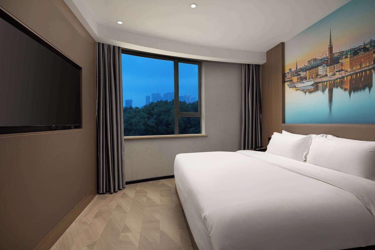 A hotel room with a large bed, a TV mounted on the wall, a window with curtains, and a cityscape painting on the wall.